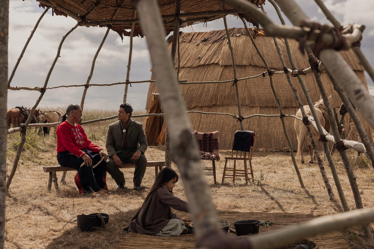 Two adults sit talking under a wooden shelter frame in a grassy field, with a child playing nearby. Behind them are traditional huts and horses, suggesting a rural or indigenous setting. The sky is cloudy.
