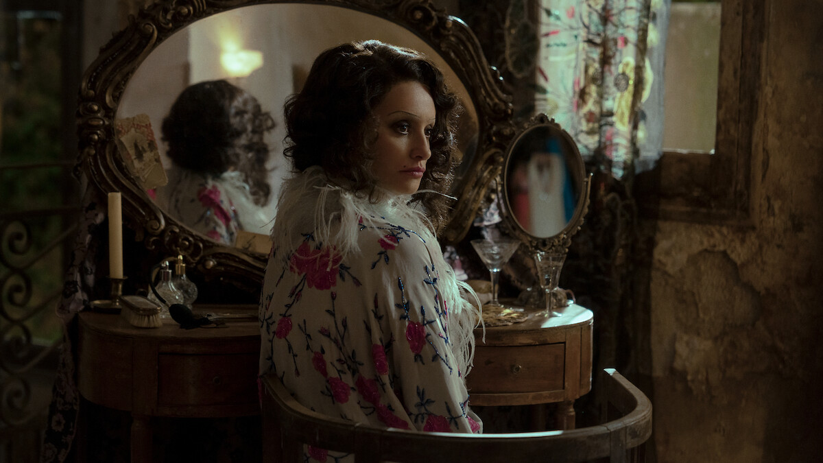 Woman with curled hair in vintage floral robe sits at ornate vanity with mirrors, old-fashioned perfume bottles, dim natural light from window, floral curtains, and weathered walls, creating an antique, moody atmosphere.