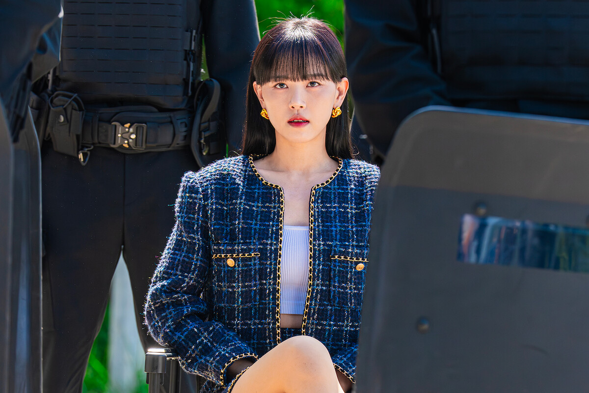 A woman in a stylish plaid jacket sits calmly among uniformed guards with shields, outdoors in bright light, creating a tense and serious atmosphere.