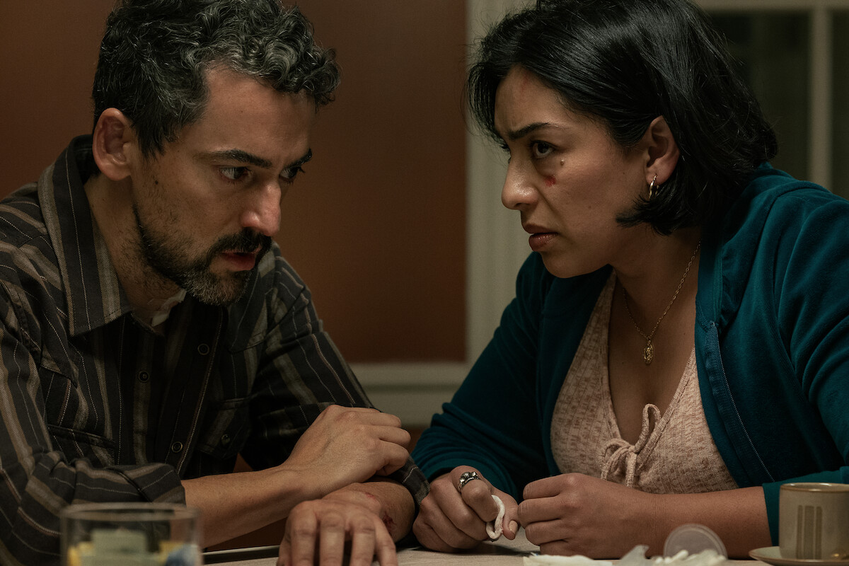 Two concerned adults sit closely at a table in a dimly lit room, engaged in a serious conversation. The setting is a domestic environment with a warm-toned wall and household items present, suggesting an intense or emotional moment.