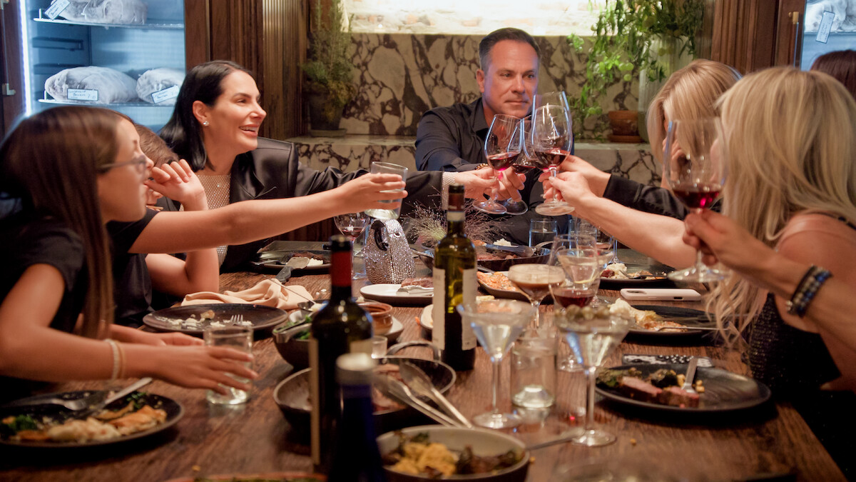 Group of people enjoying a celebratory dinner at a restaurant, raising their glasses for a toast around a wooden table set with plates of food, drinks, and wine bottles in an elegant, warmly lit indoor setting.