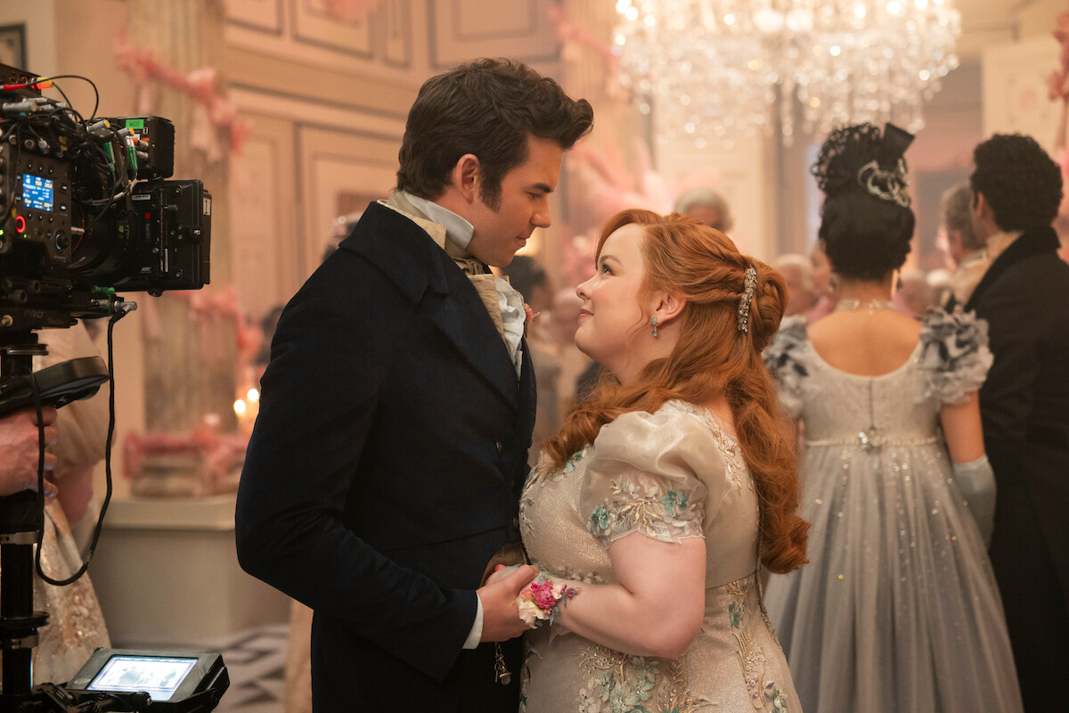 Um casal com figurinos de época dança bem próximo em um elegante salão de baile, cercado por pessoas em trajes de gala, com uma câmera de filmagem no set.