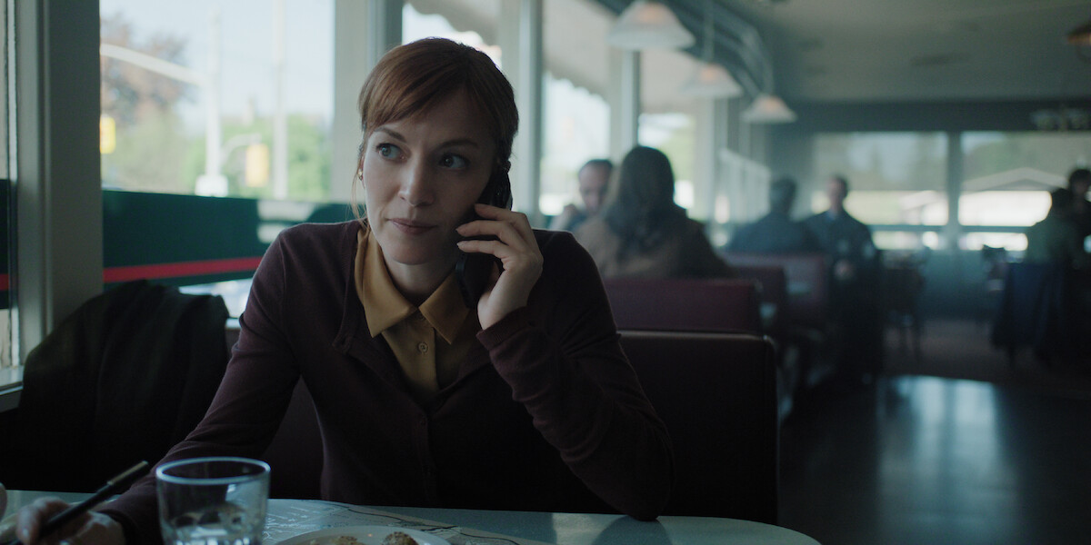 A woman with red hair sits in a diner booth talking on her phone, with a glass of water and a plate of food on the table; the diner is softly lit with other patrons in the background.