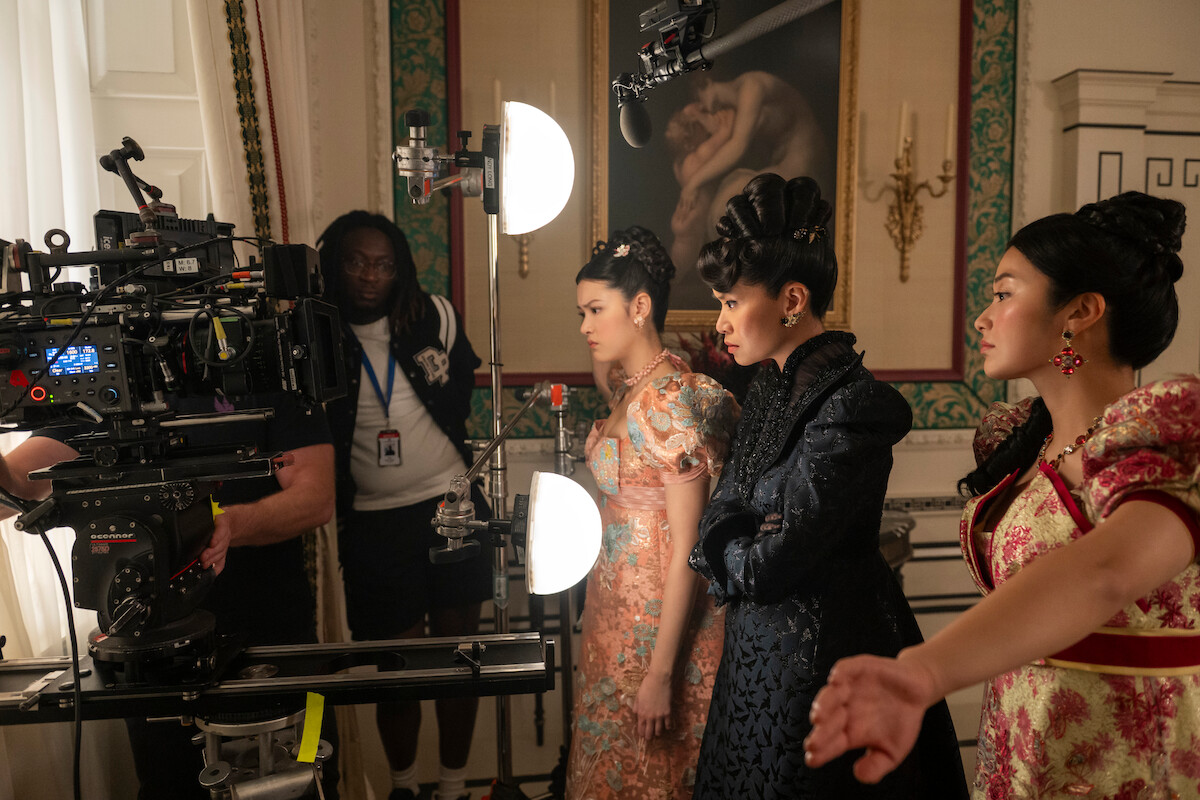 Três mulheres com figurinos de época ornamentados aguardam em um set de filmagem, de frente para uma câmera profissional e a equipe, com luzes e equipamentos de estúdio em uma sala histórica elegante, decorada com quadros e lustres.