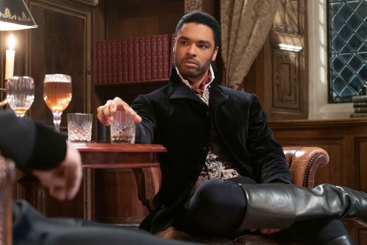 Man in period costume sits in ornate library, holding a glass at a wooden table with drinks and another person. Warm candlelight and bookshelves create an elegant, historical atmosphere.