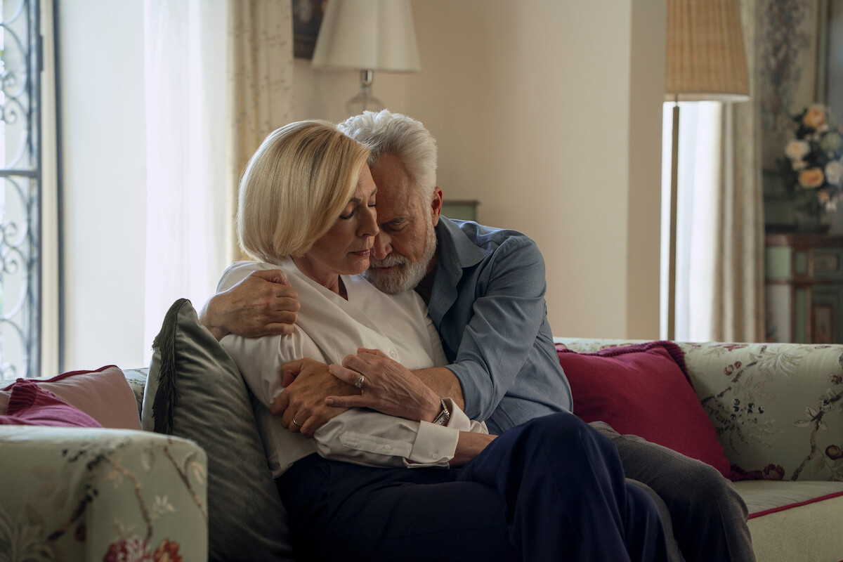 Older couple sitting closely on a sofa in a cozy living room, embracing each other with affection. The room features soft lighting, floral decor, and large windows with natural daylight.