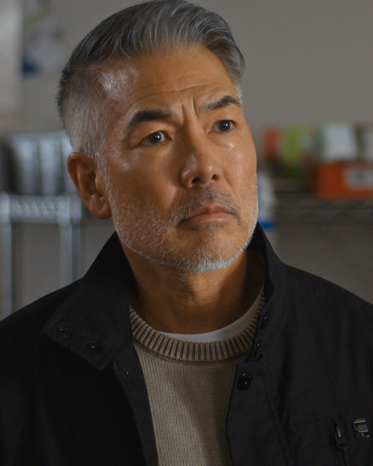 Middle-aged man with gray hair and beard wearing a black jacket and beige sweater, standing indoors in front of metal shelves and blurred kitchen supplies, looking thoughtful.