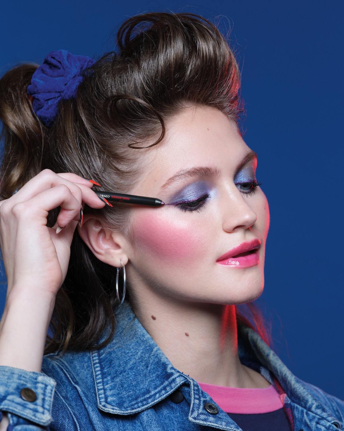 Woman with dramatic ’80s makeup, blue eyeshadow, pink blush, and glossy lips applies eyeliner. She wears a denim jacket, hoop earrings, and a blue scrunchie, set against a bold blue background with vibrant, confident mood.