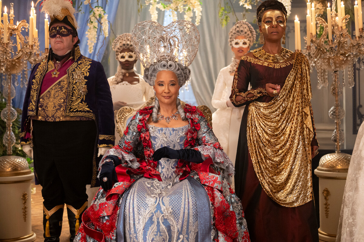 A regal woman in an ornate gown and crown sits on a throne, surrounded by elaborately dressed and masked individuals in a grand, candlelit ballroom decorated with flowers and drapes.