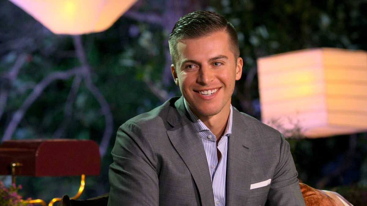 A smiling man in a gray suit sits outdoors at night, surrounded by soft lantern lighting and foliage, creating a warm and relaxed atmosphere.