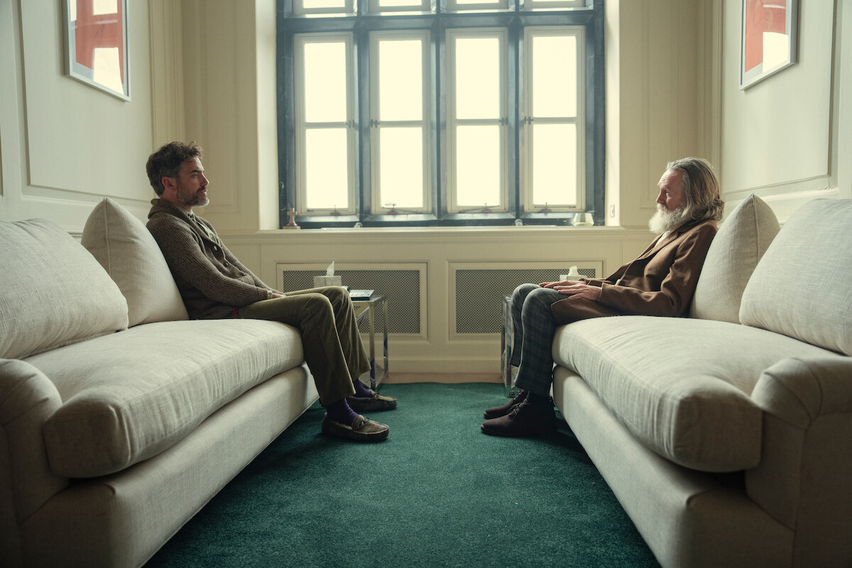Two men sitting across from each other on separate couches in a softly lit room with a large window, green carpet, and neutral decor, appearing to be in a thoughtful or conversational setting.