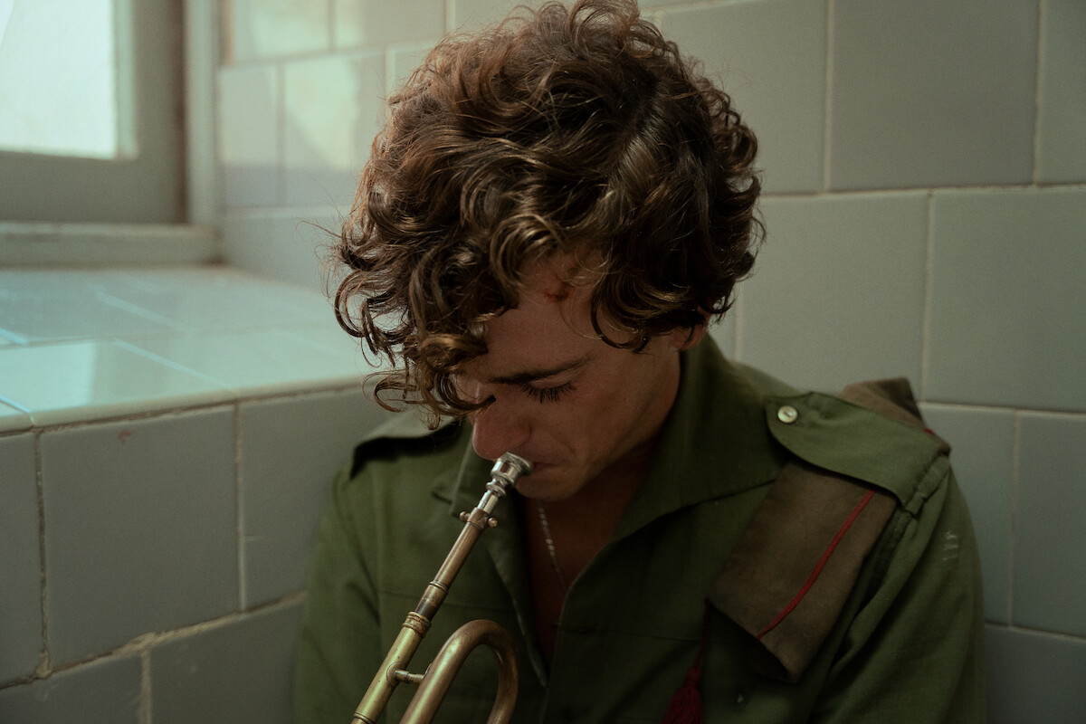 Curly-haired person in military-style green shirt sitting in tiled room by window, looking down while holding a brass trumpet, evoking a somber or reflective mood.