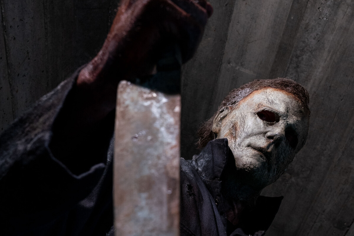 Person wearing a weathered white mask and dark clothes stands in a dim concrete environment, holding a large knife menacingly toward the camera, creating a tense and eerie atmosphere.