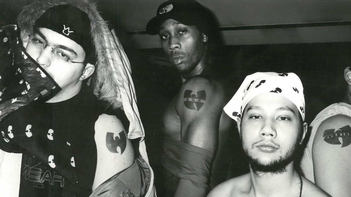 Three men pose in a black and white indoor photo, showing off Wu-Tang Clan tattoos and clothing featuring the group's logo. The environment is casual with urban streetwear and bandanas, emphasizing their affiliation and attitude.
