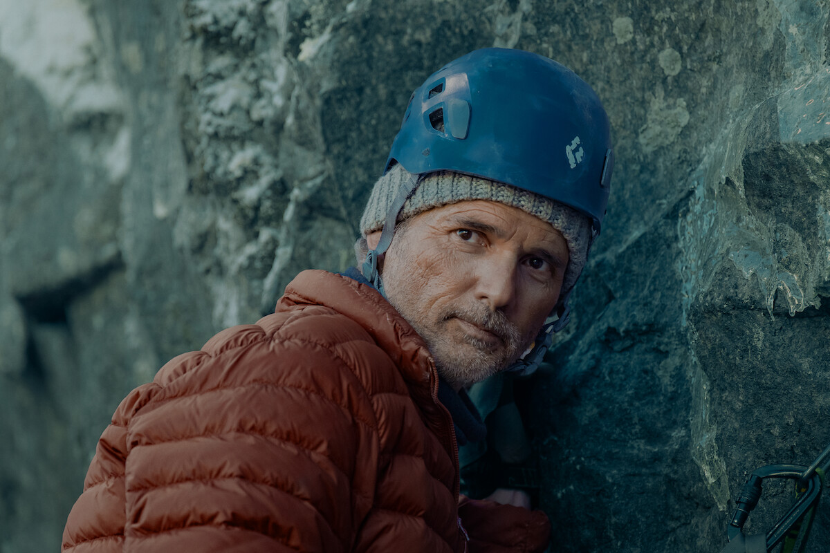 A climber in a blue helmet and red jacket rests against a rocky cliff outdoors, focused and geared for cold weather.