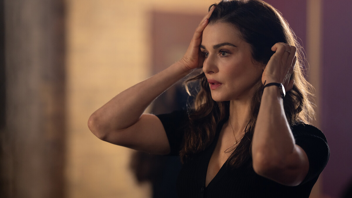A woman in a black shirt stands indoors with warm lighting, touching her head and looking concerned or thoughtful, with a blurred background suggesting an interior space.