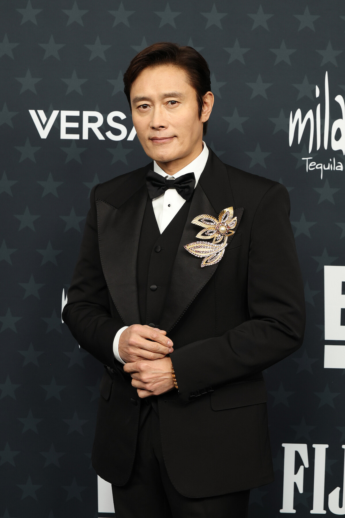 Man in a black tuxedo with a jeweled brooch stands in front of a step-and-repeat backdrop featuring logos and star patterns at a formal event or awards ceremony.