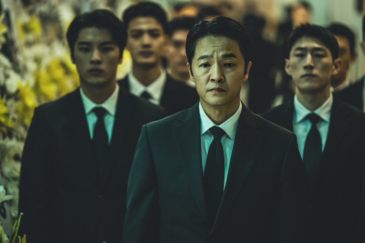 Three men stand in suits with several men in suits in the background.