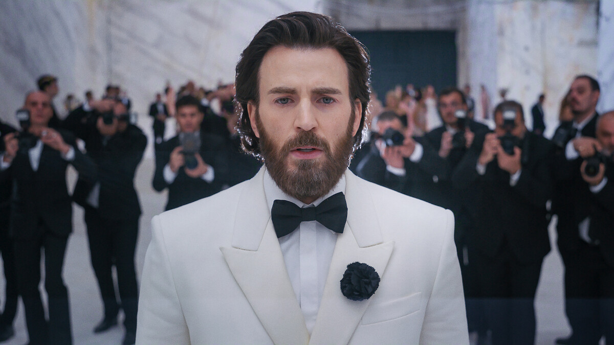 A man in a white tuxedo with a black bow tie stands in a marble hall, facing the camera, surrounded by photographers in black suits taking pictures in the background.