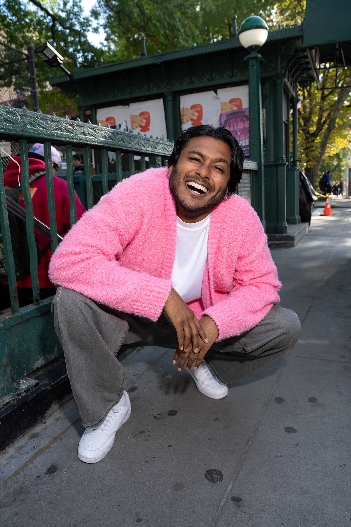 A smiling person in a pink sweater squats on a city sidewalk near a subway entrance, with sunlit trees and urban surroundings in the background.