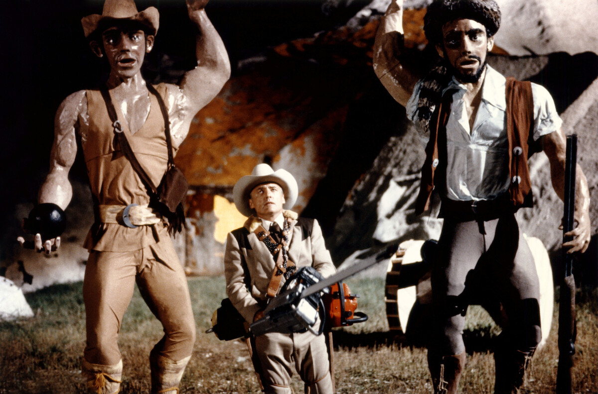 A man weilding a chainsaw among large looming cowboy doll figures
