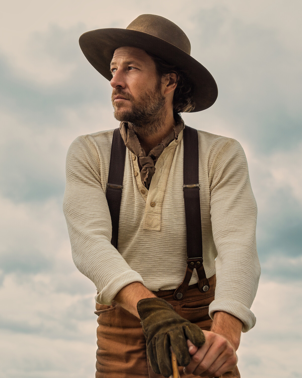 Man wearing a wide-brim hat, suspenders, gloves, and scarf stands outdoors with cloudy sky background, dressed in rustic cowboy-style clothing, gazing thoughtfully to the side.