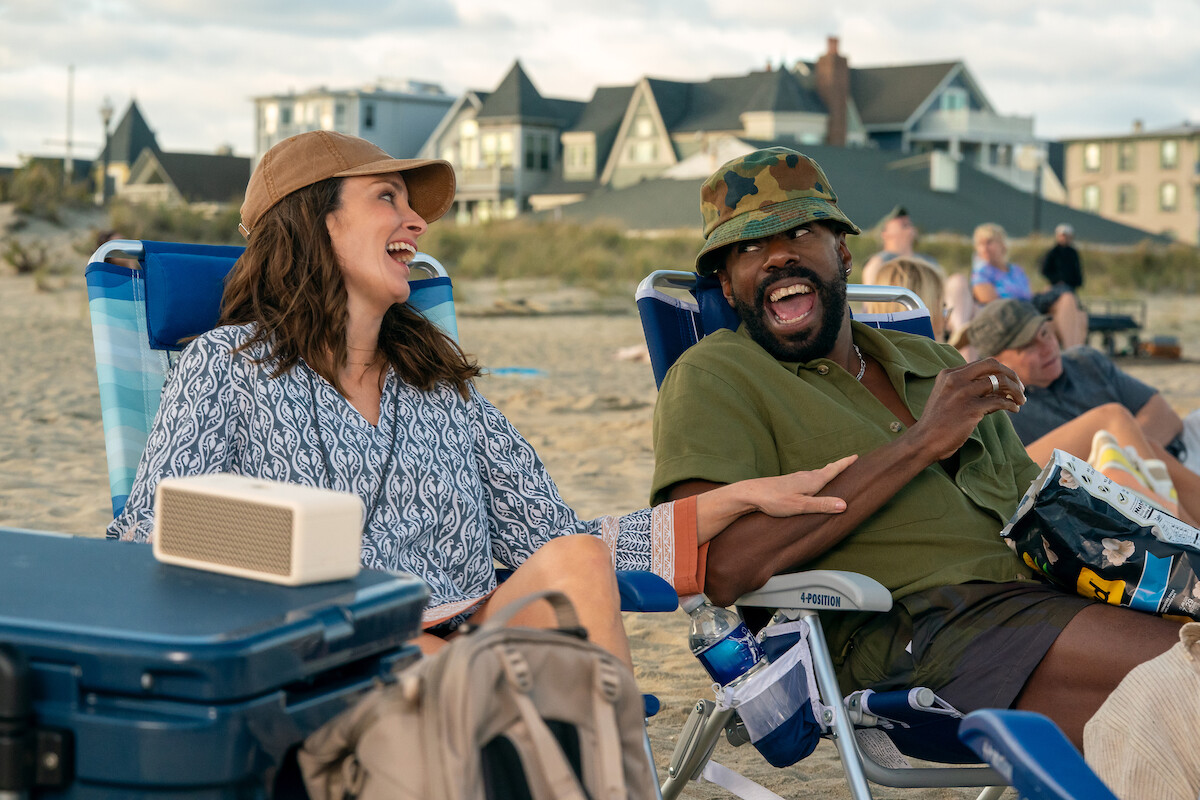 Two people relax in beach chairs, laughing together, with houses and other beach goers in the background.