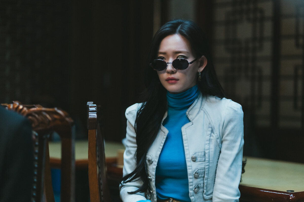 Woman with long dark hair wearing sunglasses, a blue turtleneck, and a white blazer sits indoors at a table with wooden chairs in a dimly lit room with traditional patterned wooden screens in the background.