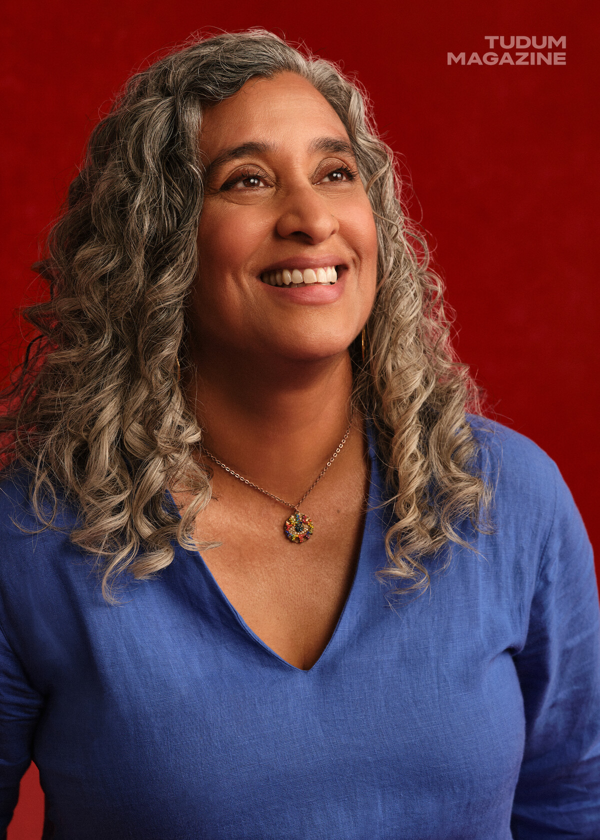 Geeta Gandbhir, director of The Perfect Neighbor, stands in front of a rich red backdrop with beautifully set curls in her graying hair, body and face at a slight angle but facing the camera while she looks slightly upwards with a happy expression on her face.