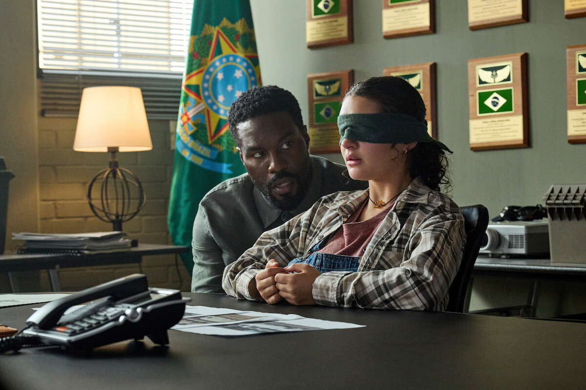 A man sits beside a blindfolded woman in an office with plaques and a flag on the wall, papers and a phone on the desk; light from a window and lamp illuminate the room.