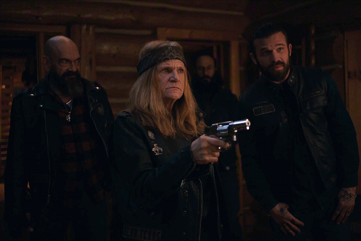 Four people in dark biker attire stand inside a dimly lit wooden cabin, one older woman in the foreground holds a silver revolver, others look on tensely, suggesting a dramatic or confrontational moment.