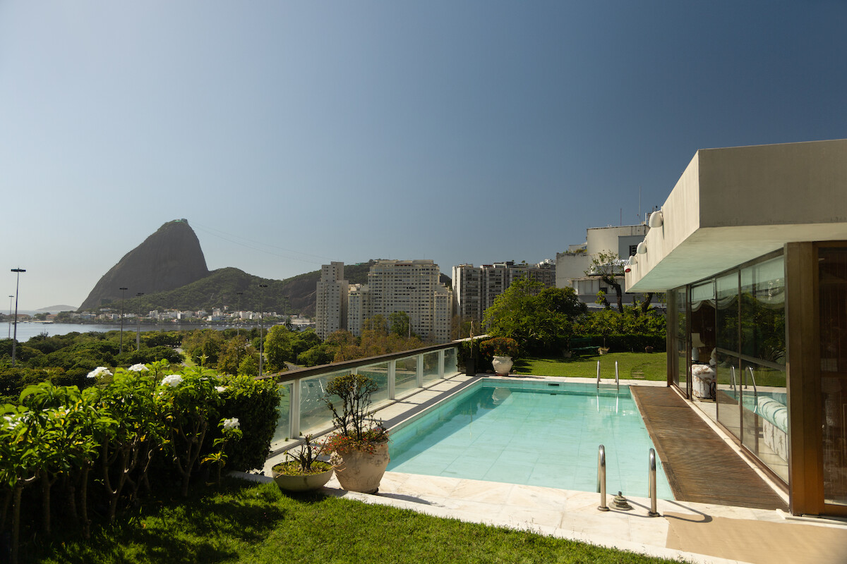 Uma área de piscina está rodeada por jardins. Ao fundo, é possível ver a zona sul do Rio de Janeiro, com o morro do Pão de Açúcar em destaque