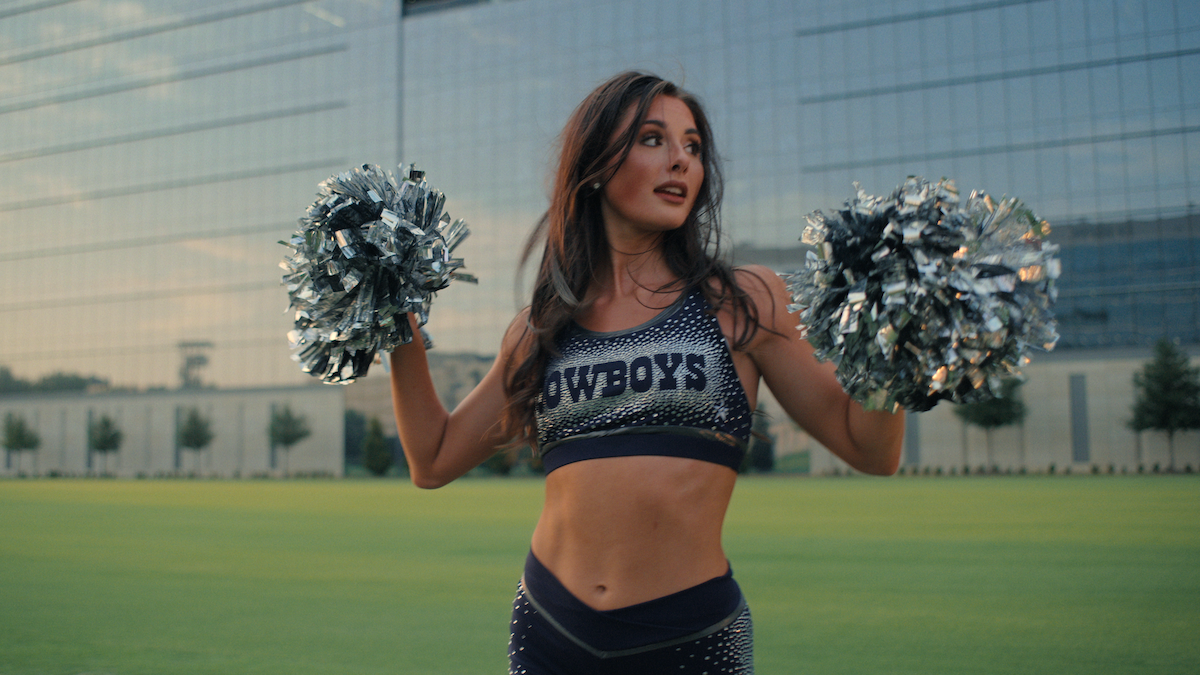 Cheerleader in a "Cowboys" uniform holding pom-poms, standing on a green field with a modern glass building in the background, looking off to the side.