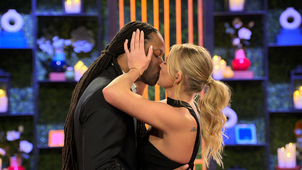 Dr. Vic St. John and Christine Hamilton kiss passionately in a romantic, candle-lit setting with colorful shelves and decorative lights in the background.