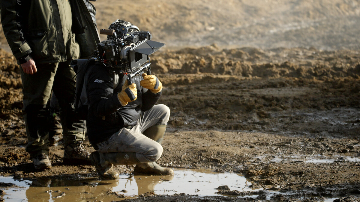 Man kneels in the mud holding a camera. 