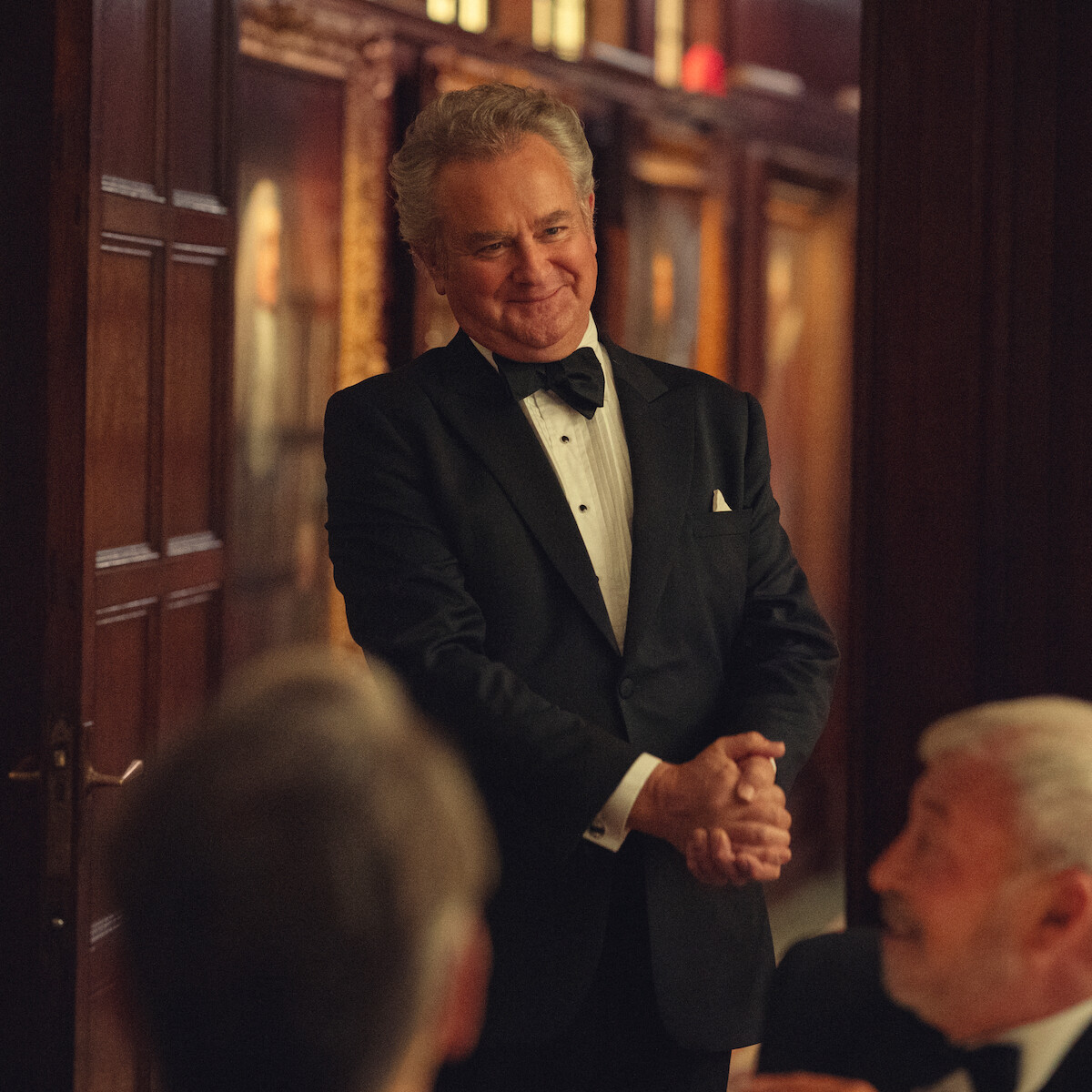 Man in a tuxedo smiling and standing in an elegant, wood-paneled room with portraits, while others in formalwear sit and interact in the foreground.