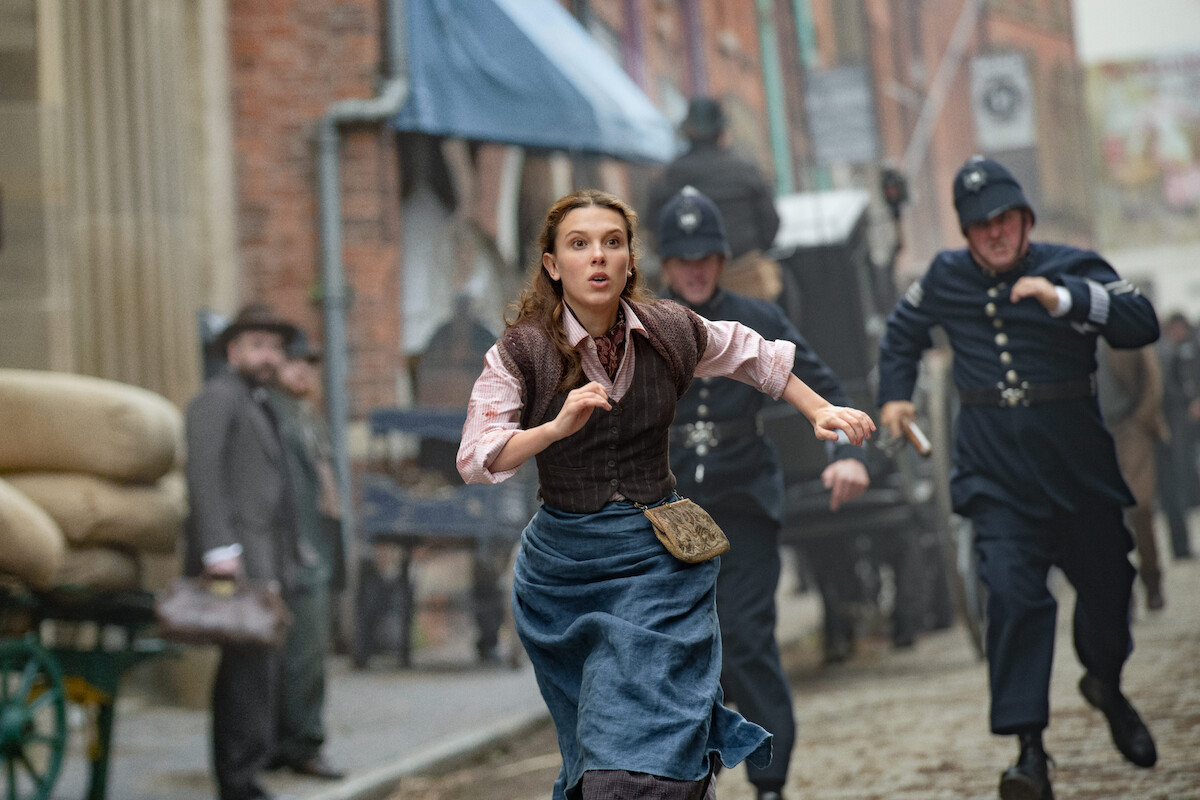 A woman in period clothing runs down an alley while a group of policemen chase her.