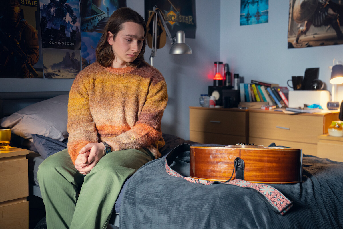 Young person in a cozy sweater sits on a bed in a neatly decorated bedroom, looking thoughtfully at a guitar on the bed; room has posters, books, warm lighting, and a calm, introspective atmosphere.