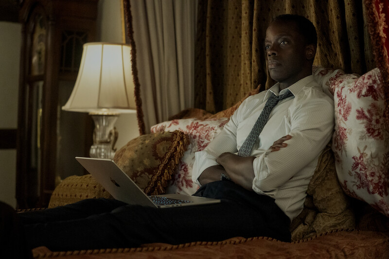 A man in a dress shirt and tie sits on a bed with his arms crossed, looking thoughtful, with a laptop open on his lap in a softly lit, ornate room.