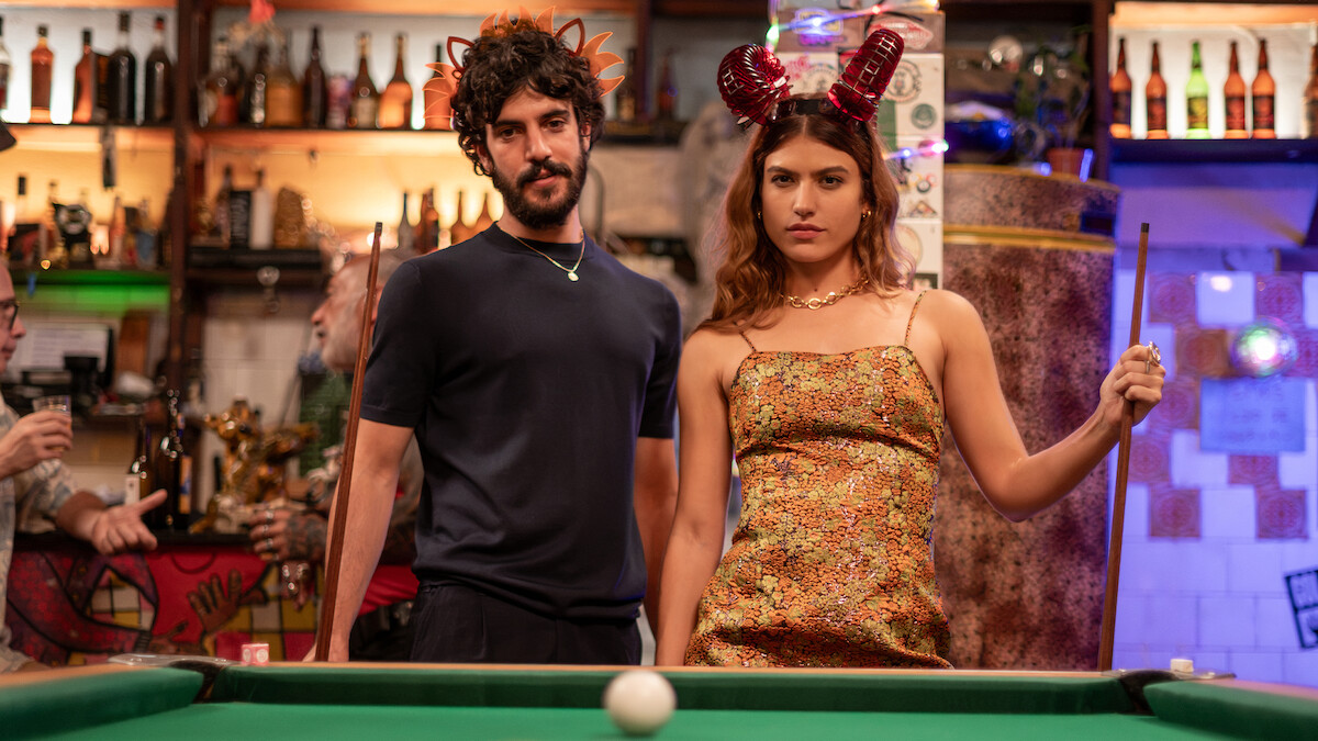 Um homem de camiseta preta, barba e cabelos grandes, e uma mulher de cabelos longos e castanhos, vestindo um vestido amarelo, se posicionam na frente de uma mesa de sinuca com a bola branca centralizada no primeiro plano