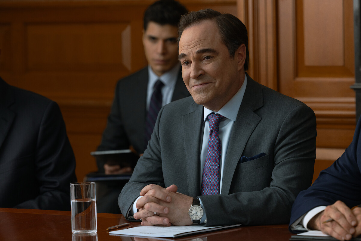 A man in a suit sits at a table in a courtroom or formal meeting room, with a glass of water, documents, and others around him, suggesting a legal or business setting. Wood paneling and another suited person are in the background.