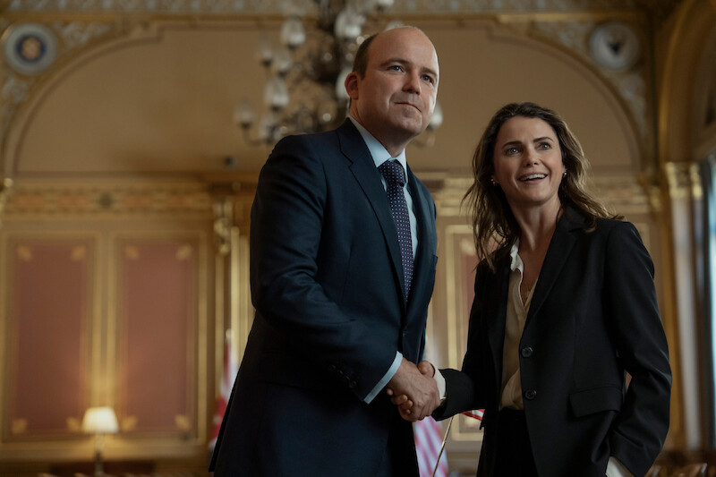 A man and a woman dressed in business attire are smiling and shaking hands in an ornate, elegant room with gold accents and chandeliers.