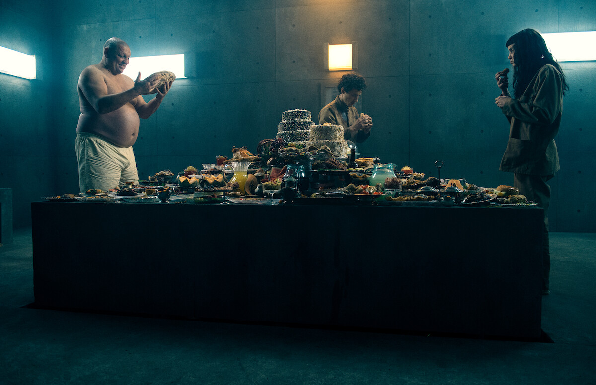 Hovik Keuchkerian as Zamiatin stands across from Milena Smit as Perempuán as the two eat from a full table of food in an image from the film ‘The Platform 2.’