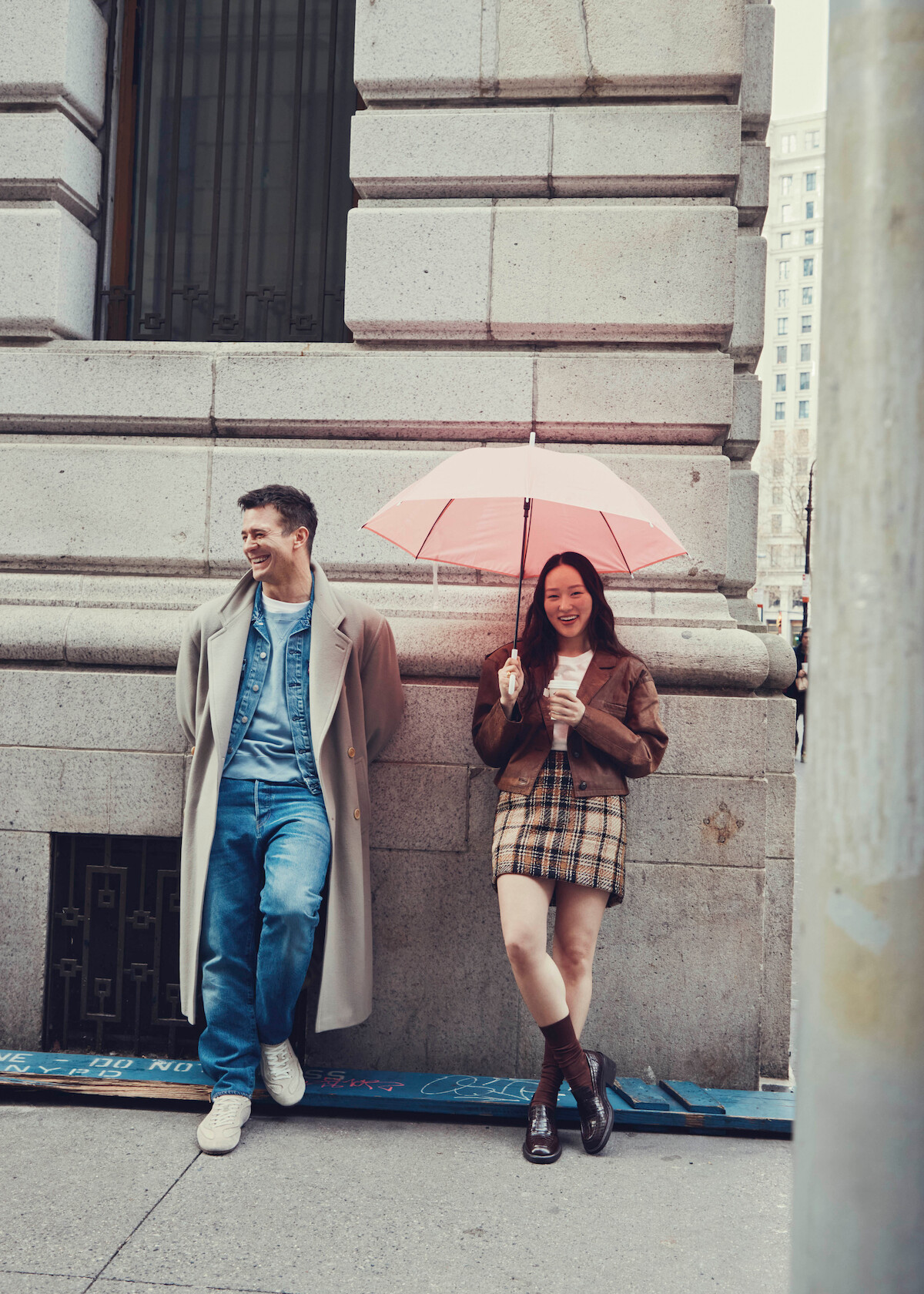 Um homem de casaco e uma mulher segurando um guarda-chuva rosa e uma bebida sorriem apoiados em um prédio de pedra em um cenário urbano, transmitindo um ambiente descontraído.