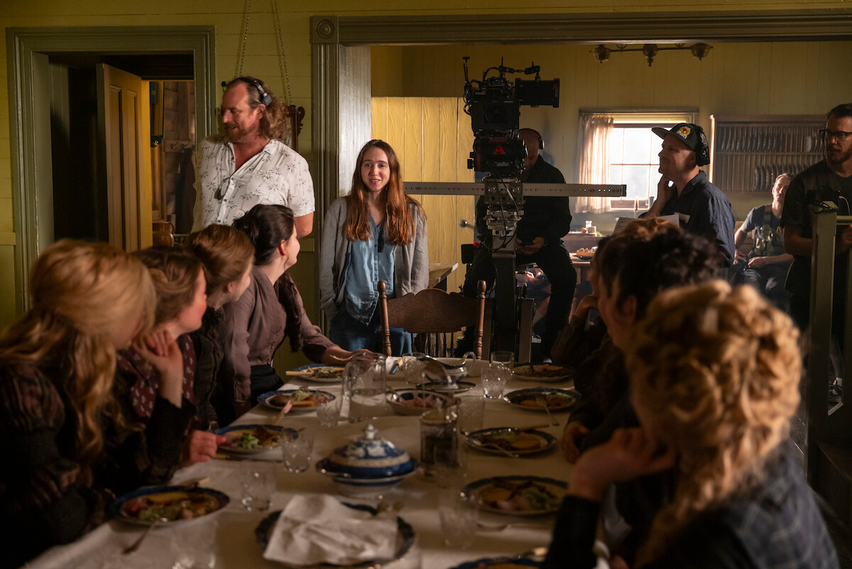 Film crew and actors gather around a dining table set with food on a film set, with cameras and lights visible, capturing a period drama scene in a warmly lit room.