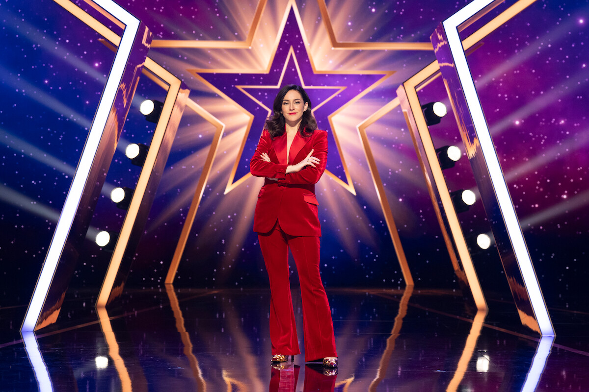 Janine Harouni stands confidently on a shiny stage with a large illuminated star and spotlights in the background, surrounded by vibrant purple and gold lighting effects.