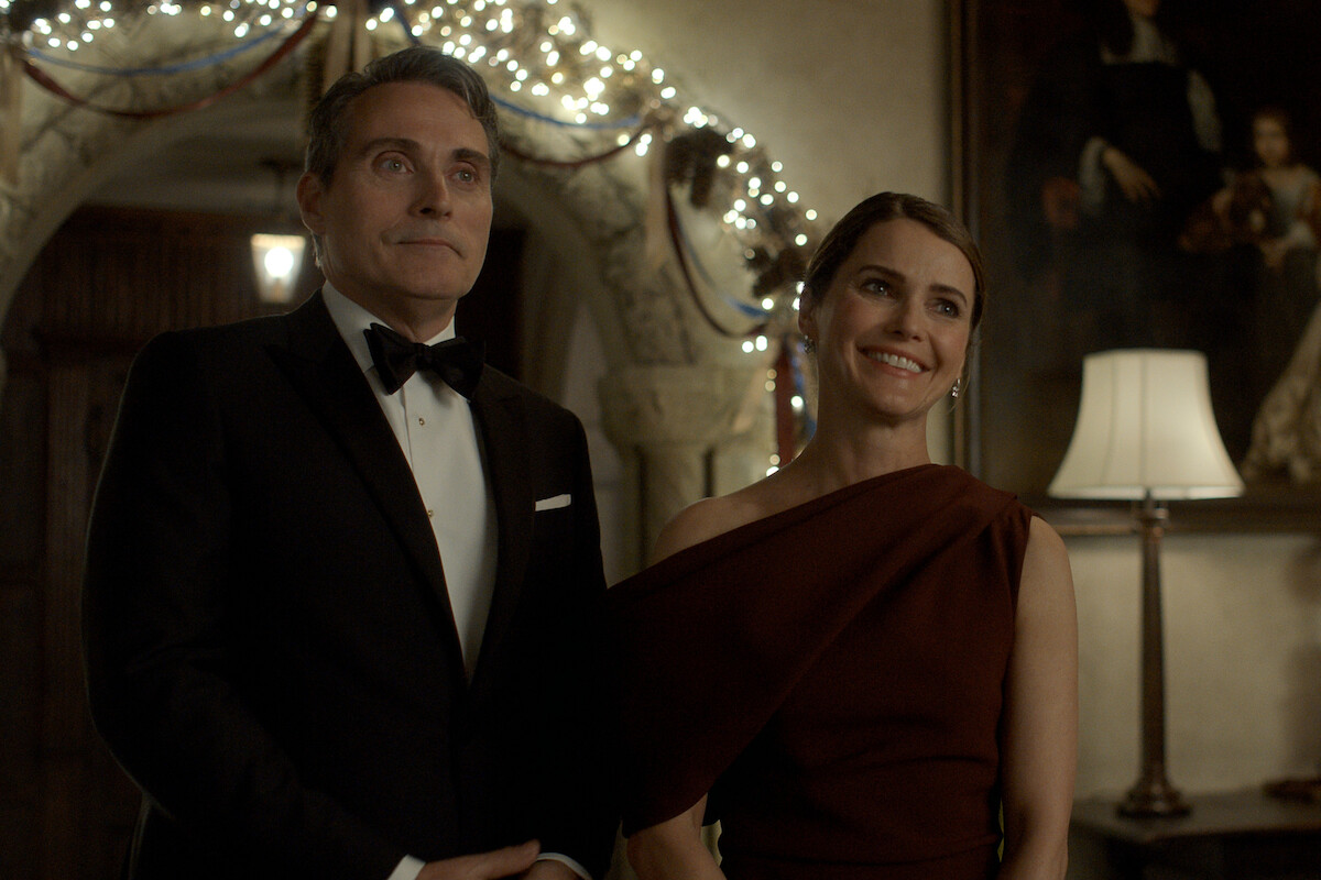 Rufus Sewell as Hal Wyler and Keri Russell as Kate Wyler stand next to each other in formal clothing while attending a party.
