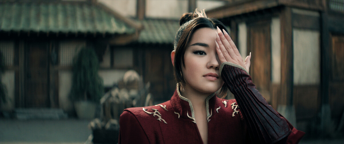 Elizabeth Yu as Azula in a red embroidered outfit stands outdoors in a traditional East Asian courtyard, covering one eye with her hand, with wooden buildings and potted plants in the background.