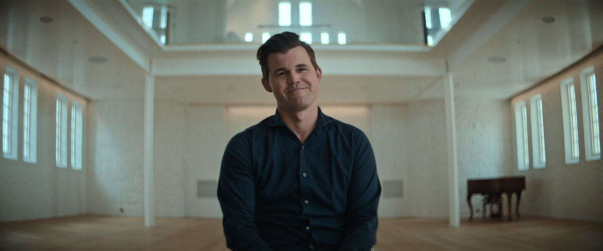 Magnus Carlsen in a dark shirt sits smiling in a spacious, bright room with wooden floors, white walls, many windows, and a piano in the background.