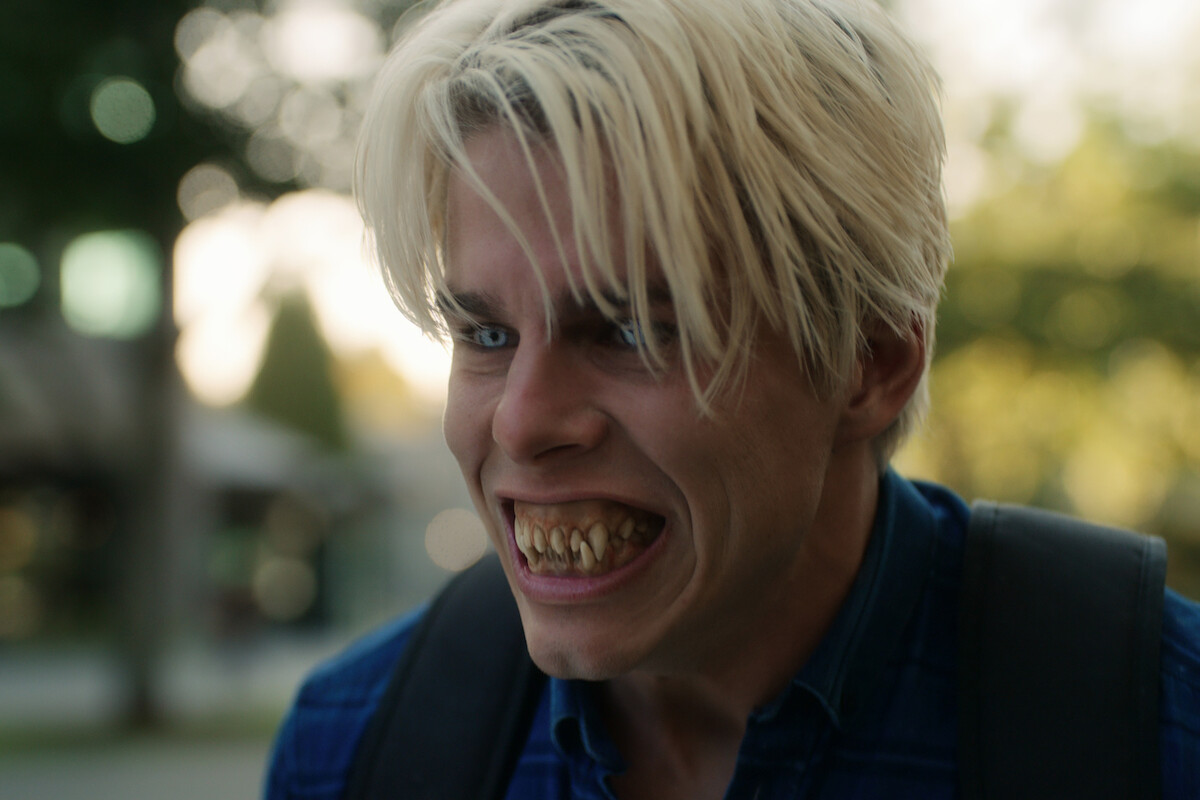 A young man with messy blond hair shows sharp, yellowed teeth and a menacing expression outdoors in a blurred park or suburban setting, wearing a blue shirt and backpack.